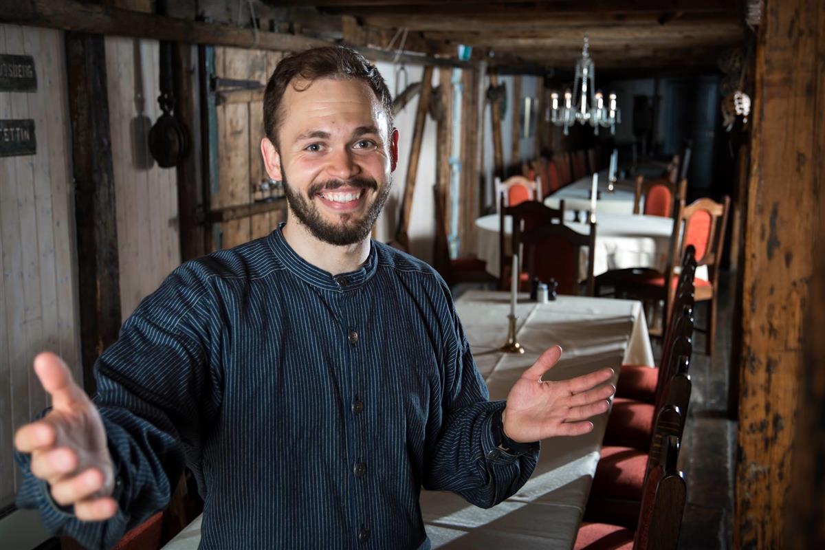 En mann smiler og ønsker velkommen inn i et lokale. - Klikk for stort bilde